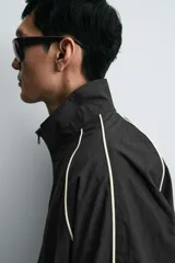 Chaqueta técnica de corte relajado en color verde oscuro, con cuello subido, cierre frontal de cremallera y detalles de líneas en contraste. Presenta bolsillos frontales con cremallera oculta y acabados elásticos en puños y ruedo.