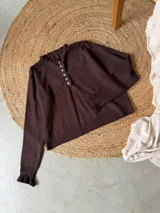 Sweater de punto color marrón chocolate, con cuello redondo y cierre frontal parcial con botones. Presenta mangas largas con puños acampanados y detalle de volados.