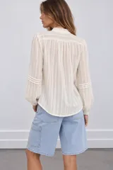 Blusa de tejido texturizado color blanco, con cuello alto con volados y cordón para atar, cierre frontal de botones y mangas largas abullonadas con detalles de encaje y tablas verticales.