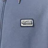 Campera canguro de felpa azul con capucha ajustable, cierre frontal completo y bolsillos tipo kiko. Presenta un parche estilo vintage en el pecho.