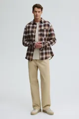 Camisa de franela de manga larga con cuello inglés, tapeta lisa y canesú en la espalda. Presenta un bolsillo superior abierto y bajo redondeado. El diseño es a cuadros en tonos marrones, beige y blanco.