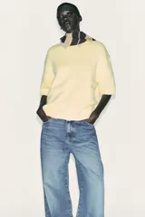 Jersey de punto color amarillo pálido, con cuello redondo y mangas cortas. Presenta un calce oversize y aberturas caladas en los laterales inferiores.