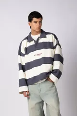 Buzo tipo polo de calce boxy con diseño de franjas horizontales anchas en color gris oscuro y blanco. Presenta cuello camisero con cierre de botones oculto y logo estampado en el pecho.