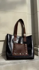 Bolso tote negro con textura símil cuero de cocodrilo y bolsillo frontal marrón con tachas plateadas. Tiene doble asa de hombro marrón y cierre superior.