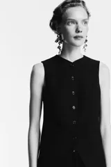 Chaleco largo negro, sin mangas, con cuello en V y cierre frontal de botones. Presenta bolsillos delanteros ribeteados y abertura en la parte trasera.