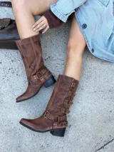 Bota biker de caña alta confeccionada en cuero vacuno color marrón con efecto desgastado. Presenta punta almendrada, taco cuadrado de madera y múltiples tiras decorativas con hebillas metálicas laterales.