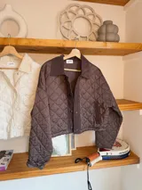 Chaqueta corta de color marrón con diseño acolchado de rombos, cuello camisero y cierre frontal con botones a presión.