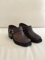 Zueco de cuero marrón con textura envejecida, diseño de punta cuadrada y taco bajo. Presenta una tira trasera con hebilla metálica y detalles de tachas plateadas.