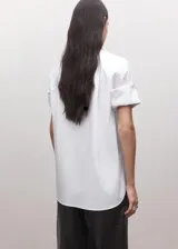 Camisa blanca de algodón con diseño recto, cuello camisero, manga corta y cierre frontal de botones ocultos con tapeta.