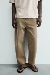 Jeans de hombre, tiro medio, color marrón claro o caqui, con corte relaxed fit y perneras anchas. Presenta cinco bolsillos y costuras laterales giradas hacia el frente.