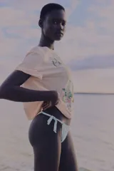 Remera de algodón color rosa pálido, corte holgado, con cuello redondo y estampado frontal de temática hawaiana con palmera, dos loros y texto que dice "HAWAII HANALEI BAY SURFERS CLUB life on the waves".