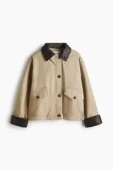 Campera levemente acolchada en lona de algodón gruesa color beige, con cuello y puños vueltos de efecto cuero marrón oscuro. Presenta cierre frontal con botones, solapa rompevientos, bolsillos delanteros de parche con solapa y bolsillos calientamanos.
