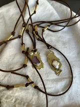 Collar artesanal con dije de base metálica irregular y piedra central roja. Presenta una estructura de tiento de gamuza marrón con detalles de tubos metálicos dorados.