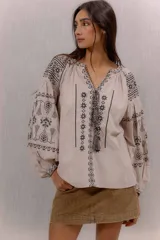 Blusa beige de estilo bohemio, con mangas largas abullonadas y cuello en V con cordones y borlas colgantes. Cuenta con elaborados bordados en contraste de color negro en el frente y las mangas.