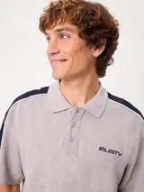 Remera polo gris melange de ajuste holgado, con cuello y cierre frontal de botones. Presenta detalles en azul marino y blanco en los hombros y mangas, y el logo de la marca bordado en el pecho.