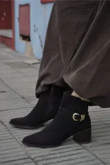 Bota corta de cuero color marrón, con taco cuadrado de madera y punta fina. Presenta una tira decorativa con hebilla metálica dorada alrededor del tobillo.