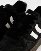 Championes urbanos Coolway modelo Goal, color negro con detalles en blanco y marrón. Confeccionados en cuero suave con puntera reforzada, cordones planos de algodón bicolor intercambiables y suela flexible de goma y caucho.