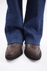 Botín de cuero vacuno color marrón, con punta fina y detalle de tachas metálicas en la parte superior. Presenta una suela de plataforma plana de color negro.