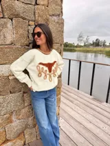 Sweater de punto fino color crema, con cuello redondo en tono contraste. Presenta un diseño frontal artesanal con figura de búfalo en cuero y detalles bordados en relieve.