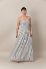 Vestido largo de tul bordado color gris claro, con lentejuelas plateadas y doradas. Modelo strapless drapeado y falda con movimiento.
