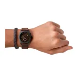 Set de reloj Fossil con caja de acero inoxidable en color negro y esfera marrón, acompañado de una pulsera trenzada de cuero en tonos marrón y negro.
