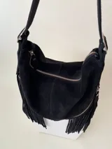 Bolso tipo hobo color tostado, hecho de cuero con flecos largos en el frente y dos bolsillos con cierre. Tiene una manija regulable con hebillas metálicas.