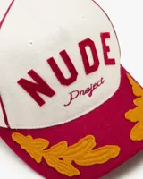 Gorra de béisbol de cinco paneles color blanca con visera color bordó. Presenta un parche frontal de fieltro con la inscripción "NUDE Project" y bordados de hojas en la visera en color amarillo mostaza.