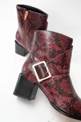Bota corta con punta afilada y taco cuadrado, confeccionada en material con textura de piel de serpiente en tonos bordó y negro. Presenta una correa decorativa con hebilla metálica dorada en el lateral.