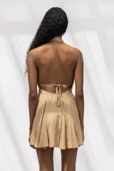 Vestido corto color beige con escote halter profundo, espalda descubierta y falda con vuelo. Presenta un diseño fluido y ligero con detalle de frunces en la cintura.
