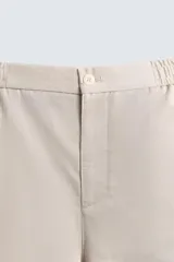 Pantalón de hombre de corte relajado, color beige claro, con cintura elástica, cierre frontal con botón y cremallera, bolsillos delanteros y bolsillos traseros de vivo.
