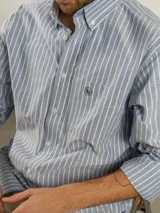 Camisa de corte regular confeccionada en tejido Oxford de algodón, con diseño de rayas verticales finas. Presenta cuello abotonado, cierre frontal con botones y un bolsillo en el pecho.
