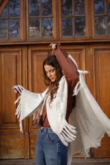 Poncho tejido de color blanco, con flecos en los bordes y detalles de bordado en crochet a lo largo de la abertura frontal. Cuenta con un cierre de cordones con borlas en el cuello.