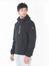 Campera rompeviento Calvin Klein de color negro, con cierre frontal, capucha ajustable y logo estampado verticalmente en el frente. Presenta puños elásticos y un parche con el logo en la manga.