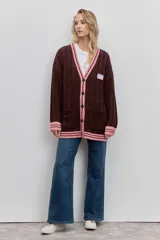 Cardigan de punto trenzado en color marrón oscuro, con detalles en rosa en el cuello en V, puños y ruedo. Presenta cierre frontal con botones, bolsillos delanteros y un parche decorativo en el pecho.