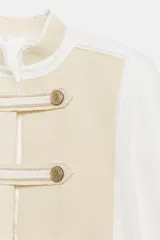 Chaqueta de estilo militar de ZARA Woman Collection, confeccionada en mezcla de algodón en color blanco y beige. Presenta cuello subido, manga larga con puños anchos en contraste y cierre frontal con alamares y botones metálicos en relieve. Tiene bajo asimétrico con abertura en la espalda.