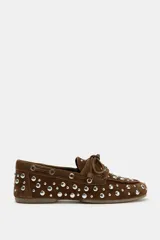 Zapato tipo náutico de serraje color marrón, con detalle de tachas metálicas plateadas en todo el cuerpo y cordones decorativos en la parte delantera. Presenta punta redonda y suela plana.