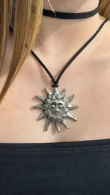 Choker compuesto por una cinta negra de gamuza y un dije metálico plateado con forma de sol con rostro.
