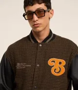 Campera bomber de estilo college con cuerpo a cuadros en tonos marrones y mangas largas de cuero sintético negro. Presenta cuello, puños y cintura acanalados a rayas, cierre abotonado, bolsillos laterales y aplique bordado de letra "B" en el pecho.