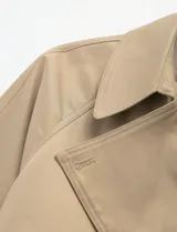 Gabardina corta de color beige, con diseño de doble botonadura, solapas anchas y cinturón ajustable en la cintura. Presenta bolsillos laterales y mangas largas.