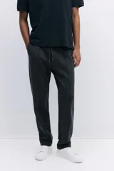 Pantalón tipo jogger de punto en tono gris oscuro, con cintura elástica y cordón ajustable. Presenta un pliegue cosido en el frente de cada pierna, bolsillos laterales y un bolsillo insertado trasero.