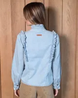 Camisa de denim celeste con cuello alto con volados, botonadura frontal y detalles de volados verticales a lo largo del frente. Cuenta con mangas largas abullonadas y puños con volados.