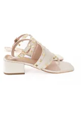 Sandalia de cuero color offwhite con taco bajo cuadrado. Presenta diseño de tira entre los dedos, correa ajustable al tobillo con hebilla y detalle de tachas metálicas doradas en las tiras.