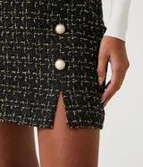 Pollera corta de tweed texturizado en color negro con hilos dorados, presenta botones decorativos con centro blanco y una pequeña abertura frontal en el ruedo.