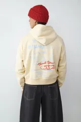 Canguro de color crema con capucha y estampado gráfico en color rojo en el pecho. Presenta mangas largas con puños elásticos y un diseño holgado.