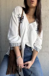 Camisa blanca de manga larga con escote en V y cierre frontal de botones. Presenta delicados bordados florales en tono oscuro en los hombros y puños.
