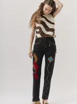 Pantalón de jean tiro alto tipo mom-fit, con parches de colores en forma geométrica, detalles de tientos de cuero trenzados en la cintura y adornos de canutillos en la parte frontal.