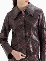 Chaqueta de cuero vacuno color marrón con acabado brillante, diseño de cuadros con líneas finas en tonos rojizos y beige, y detalle de volados de cuero aplicados en forma de trama. Posee cuello camisero, cierre frontal con botones y mangas largas.