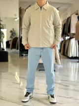 Campera de gabardina beige de corte boxy fit, con cuello camisero, cierre frontal de cremallera blanca y bolsillos laterales ribeteados.