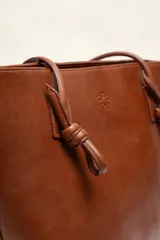Cartera tote de cuero suave color marrón, con diseño amplio y líneas simples, con tiras de hombro decoradas con nudos artesanales y pequeño logo grabado en el centro.