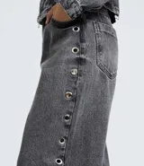 Pantalón baggy de jean color gris desteñido, con corte amplio y recto. Presenta una hilera de ojales metálicos decorativos a lo largo de los laterales y cierre frontal con botón y cremallera.